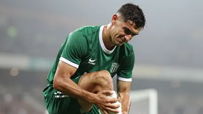 Mid-30s muscular defender, head tilted, grimacing as he clutches a swollen ankle, dark green training shirt with white...