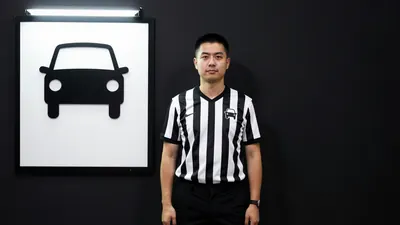 A 30-year-old male stadium operations manager, wearing a black and white striped shirt and black trousers, stands in...