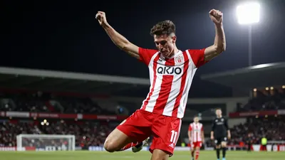 A young winger in a red and white striped kit, mid-kick, celebrating a goal with arms raised