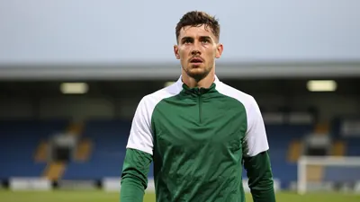 A determined 25-year-old left-back in a plain dark green and white training top, shot with an 85mm f/1.4 lens, stands on...