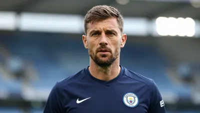 A mid-30s male coach in a navy blue Swansea City training shirt, head-and-shoulders, focused expression, blurred stadium...
