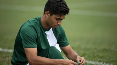 Mid-30s, athletic midfielder, dark hair, focused expression, wearing a plain dark-green shirt with a white vertical...