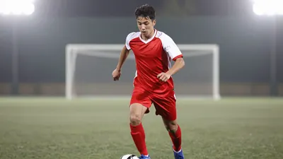 A young, athletic winger in a red and white football kit, mid-action with the ball at his feet, running forward with...